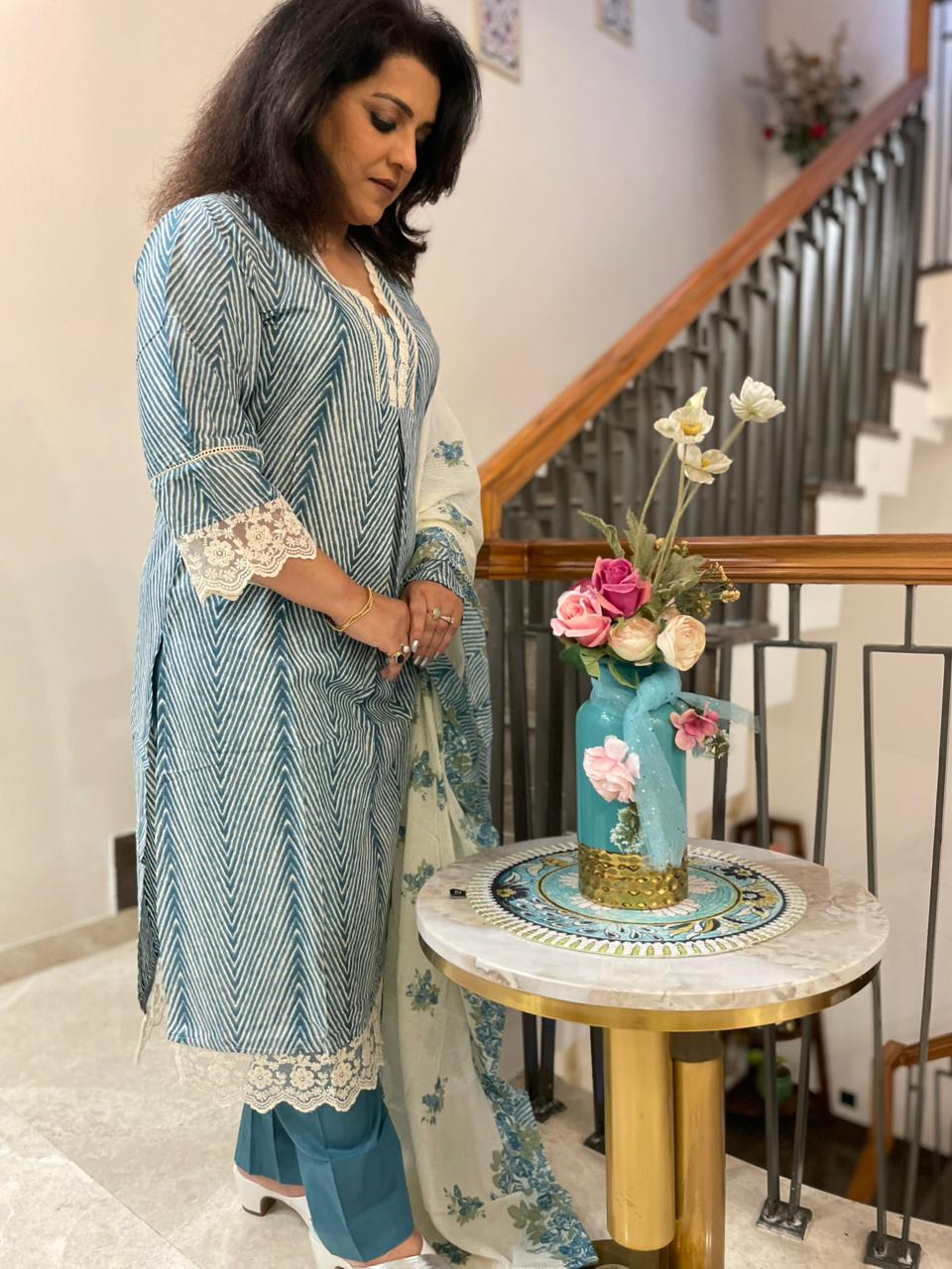 Woman in a blue and white traditional outfit standing next to a table with a decorative vase and flowers.
