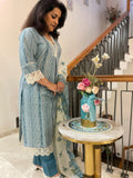 Woman in a blue and white traditional outfit standing next to a table with a decorative vase and flowers.